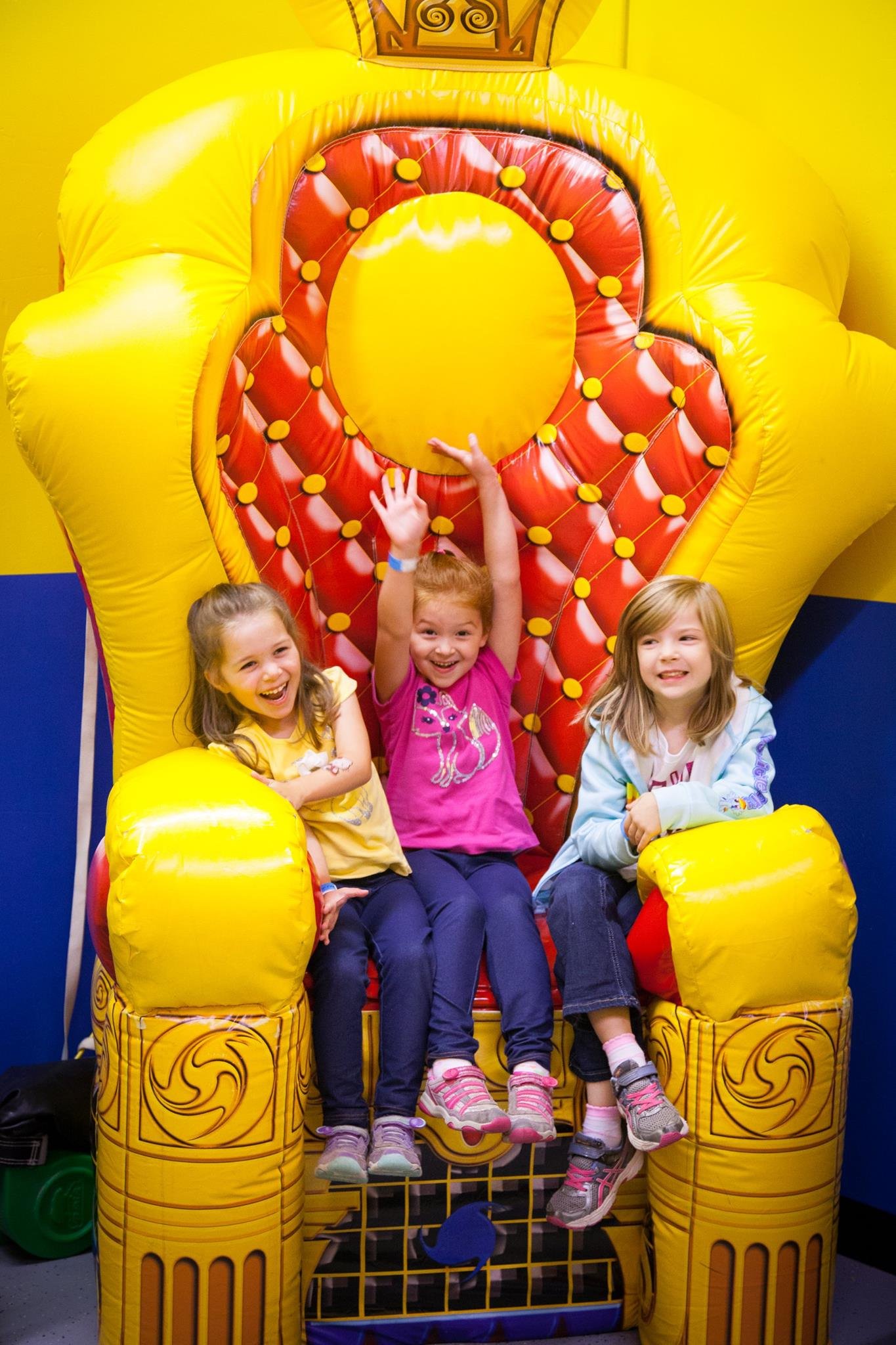 Three girls sitting and posing joyfully on a bright yellow and red inflatable throne against a colorful bouncy castle.