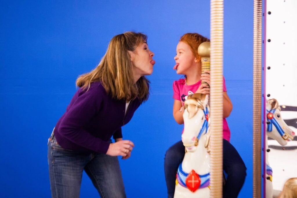 Two people are playfully sticking their tongues out while sitting on a merry-go-round horse against a blue background.