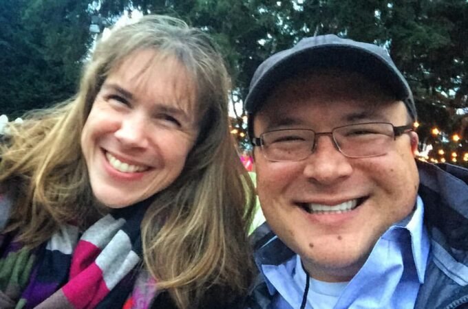 Two smiling people pose for a selfie outdoors at night with String lights and evergreen trees in the background. Close-up of smiling faces during a winter evening outing.