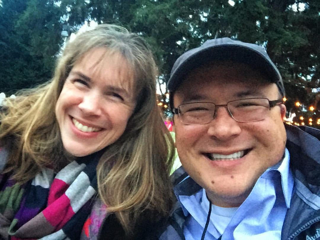 Two smiling people pose for a selfie outdoors at night with String lights and evergreen trees in the background. Close-up of smiling faces during a winter evening outing.