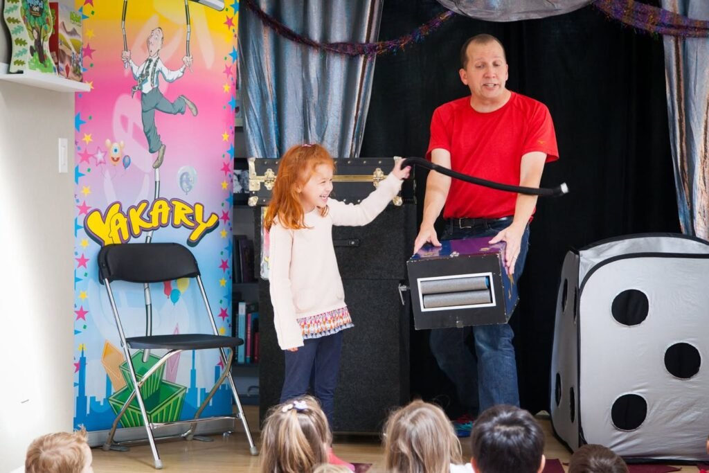 A performer and a child on stage with magic props including a large die and decorated backdrop.