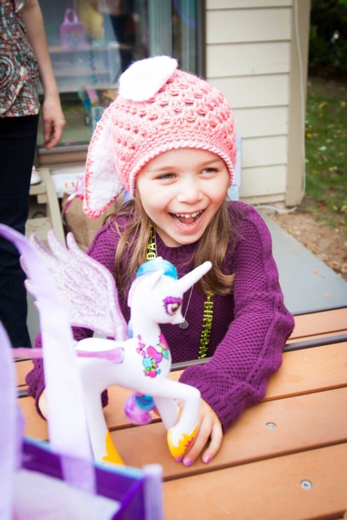 A child wearing a pink crocheted hat smiles while holding a white unicorn toy at an outdoor table, with a joyful expression matching the playful moment.