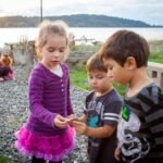 Children on a rocky lakeside path at sunset, holding small items together while a lake stretches into the distance.