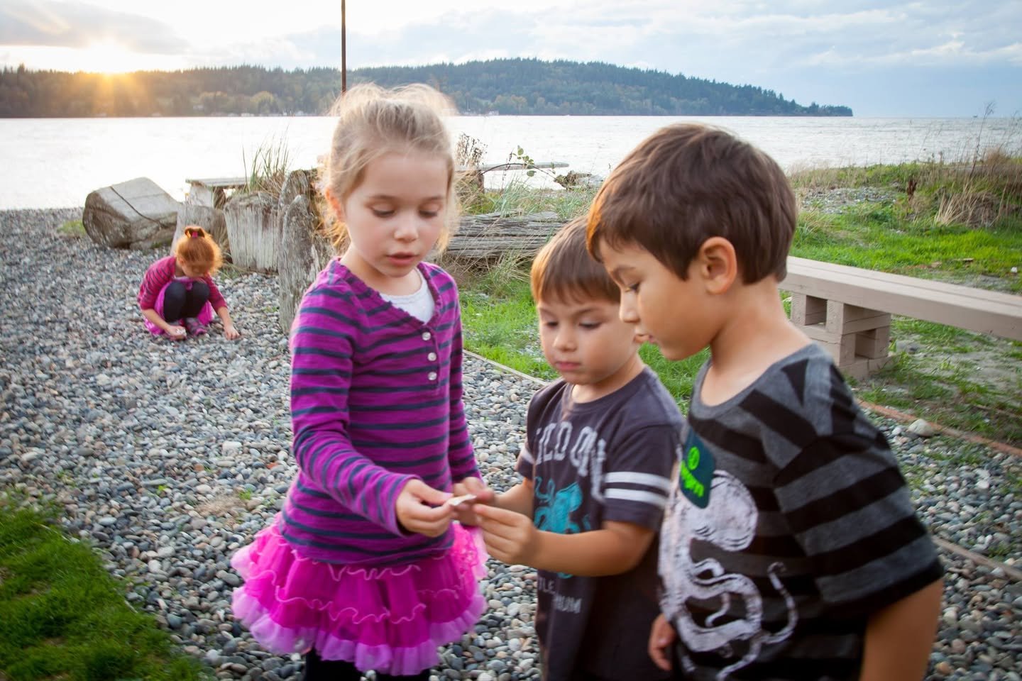 Children on a rocky lakeside path at sunset, holding small items together while a lake stretches into the distance.
