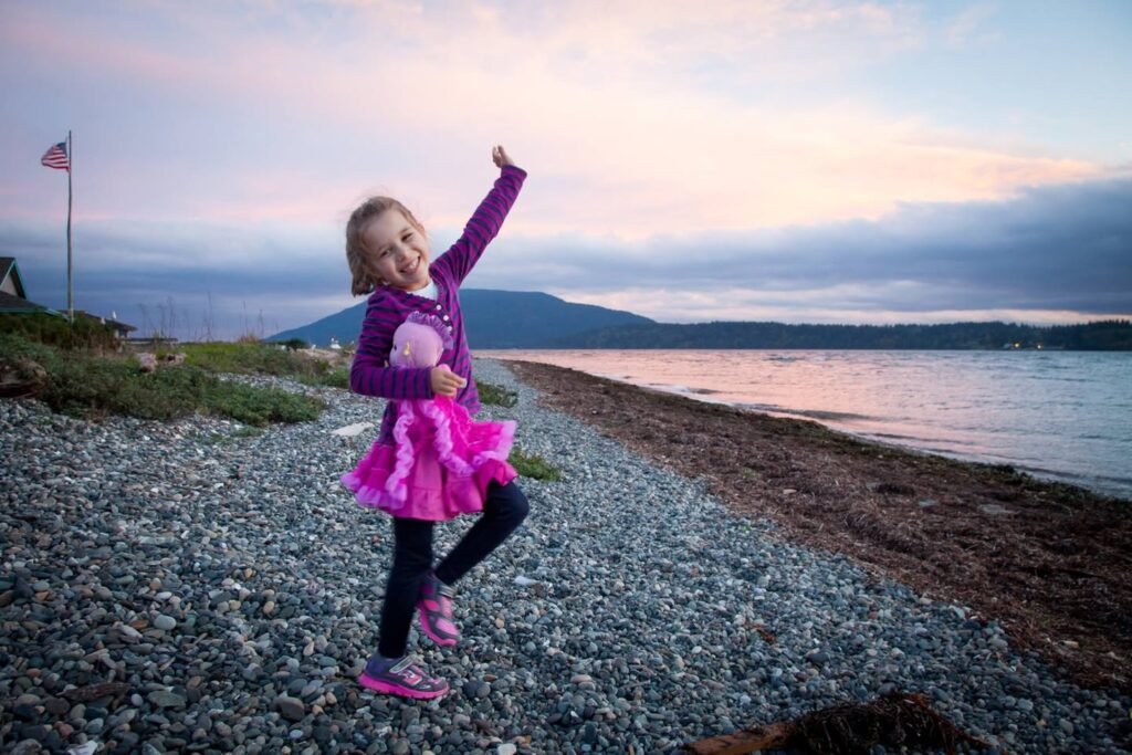 A child in bright pink plays on a gravel shoreline at dusk, with a gentle sky and distant water reflecting the blue hour. A flag flutters softly on the beach in the background.