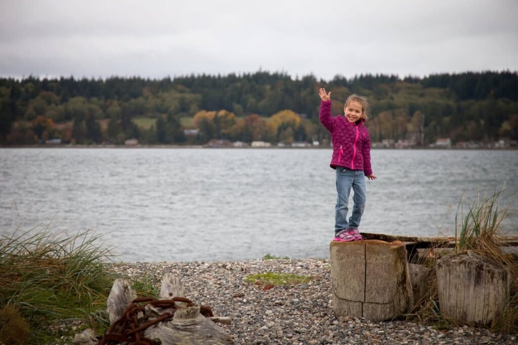 A person waving from a wooden post on a gravelly lakeside shore.