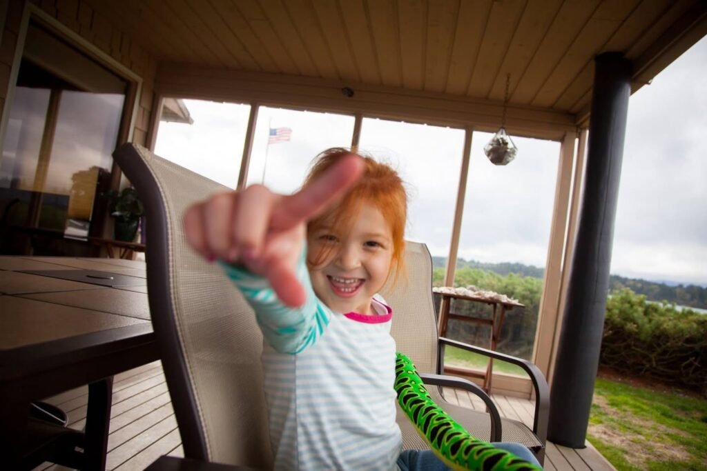 A smiling child points directly at the camera while sitting on a porch chair. The background features a lush green landscape and overcast sky.