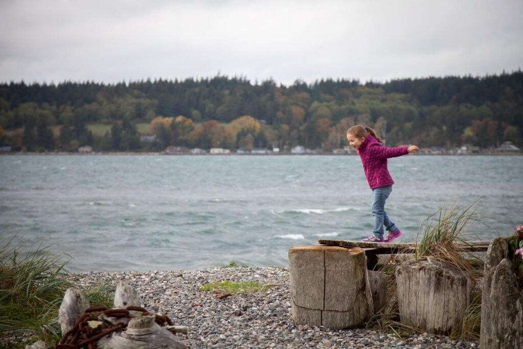 A child in a purple jacket balances on a wooden dock by the lake.