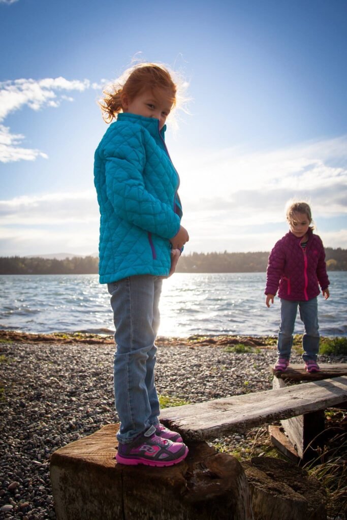 Two children wearing bright fleece jackets balance on wooden logs near a lake.