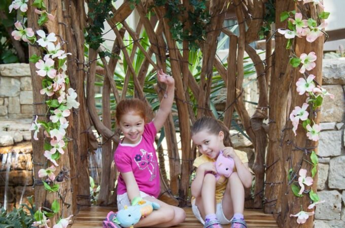 Two young children sitting on a rustic wooden bench constructed from branches, decorated with climbing pink and white flowers in a garden setting.