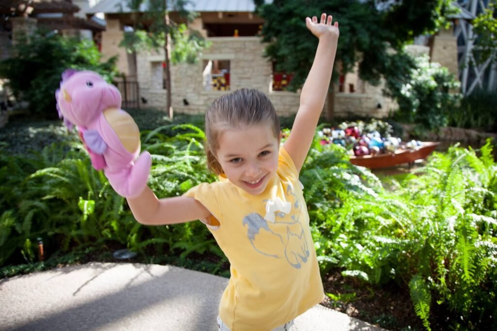 A smiling child in a yellow shirt holds a pink flamingo toy. The background is a lush green garden with a house and small boat.