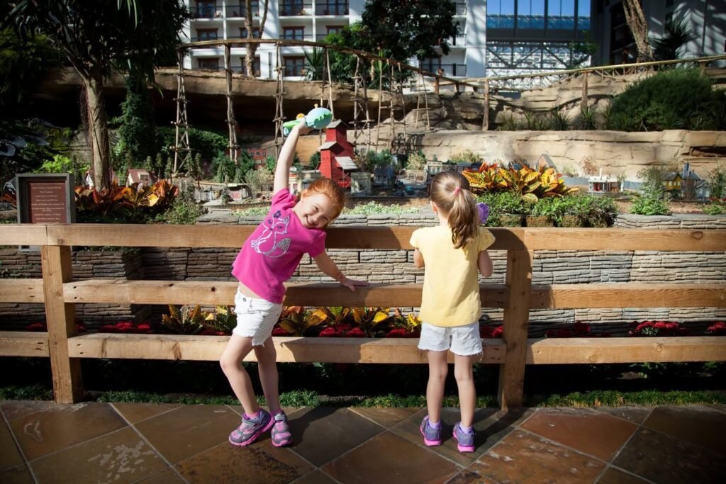 Two children playing with a toy on a stone patio in front of a wooden planter box. Lush plants and flowers are visible in the background garden area.