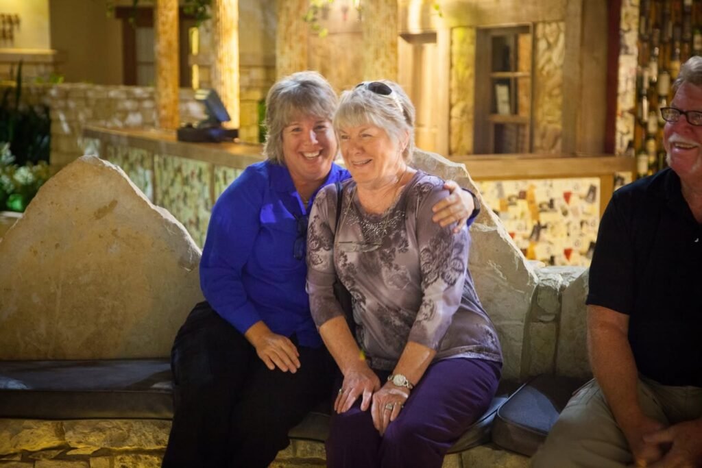 Two friends sit closely together on a couch in a cozy living room. A third person is partially visible to the side, suggesting a shared moment among good company.
