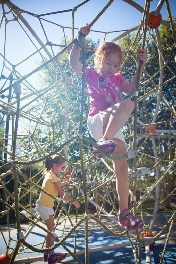 Children exploring a large rope net climbing structure at a sunny outdoor playground.