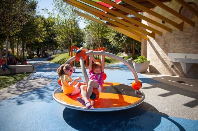 Children playing on a colorful balance disc under a wooden pergola in a sunny park. Close-up shots show trees, pathways, and the playground equipment.