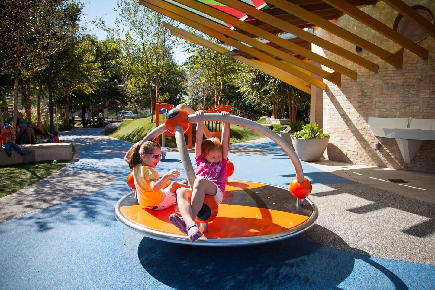 Children playing on a colorful balance disc under a wooden pergola in a sunny park. Close-up shots show trees, pathways, and the playground equipment.