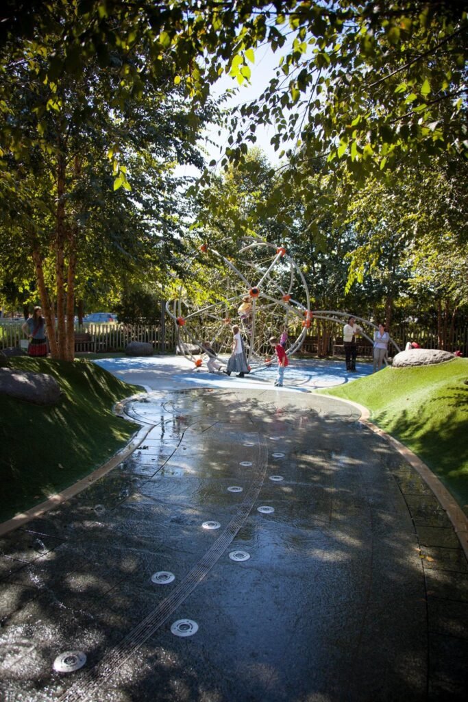 A wet walkway with circular lights leads through a garden of trees and shrubs.