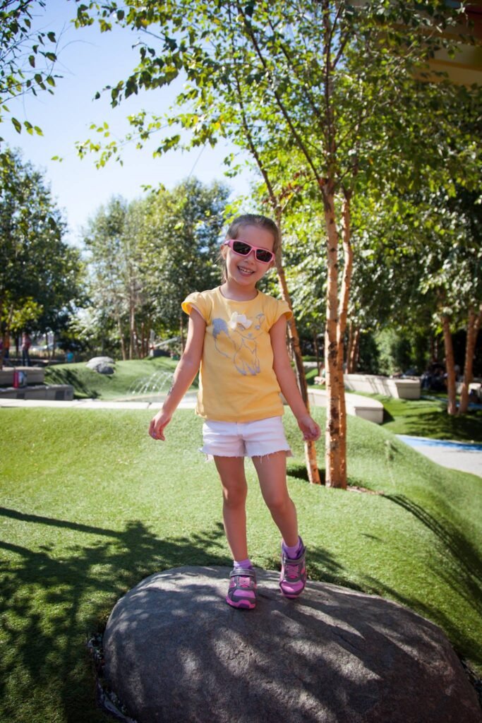 A child balances on a large rock in a lush, sunlit park.