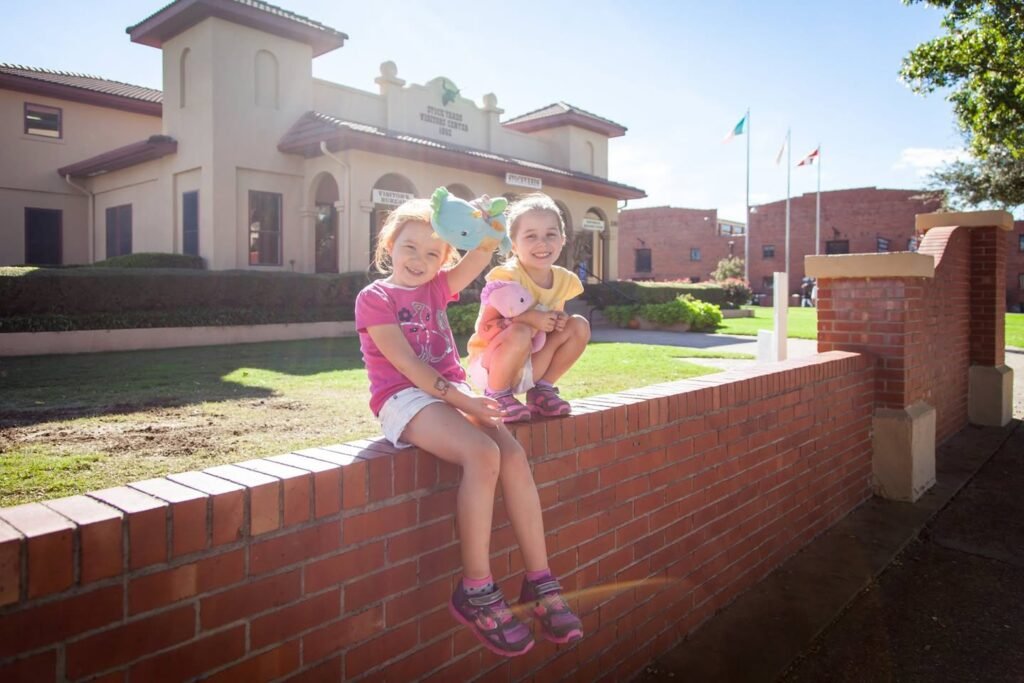 Two kids sit on a red brick wall eating ice cream outside a beige building. They hold blue and pink ice cream bars on a sunny day.