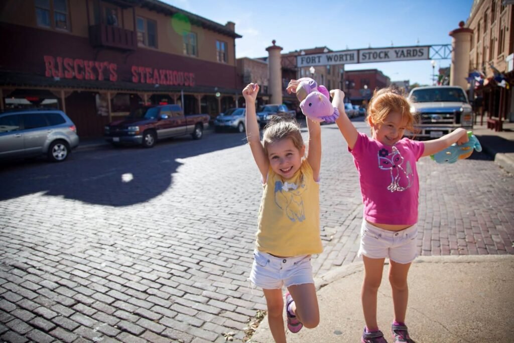 Children joyfully walking on a cobblestone street in a historic city center. The setting features vintage storefronts and a sunny evening atmosphere.