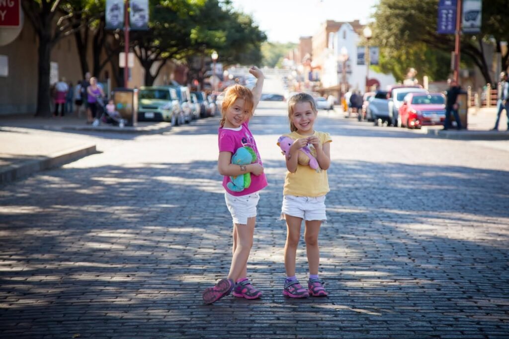 A pair of children in colorful summer clothes stands in the middle of a sunlit cobblestone street. The background features parked cars and trees along the sidewalk.