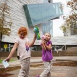 Two children with balloons walking in front of a modern building with a distinctive angular glass structure.