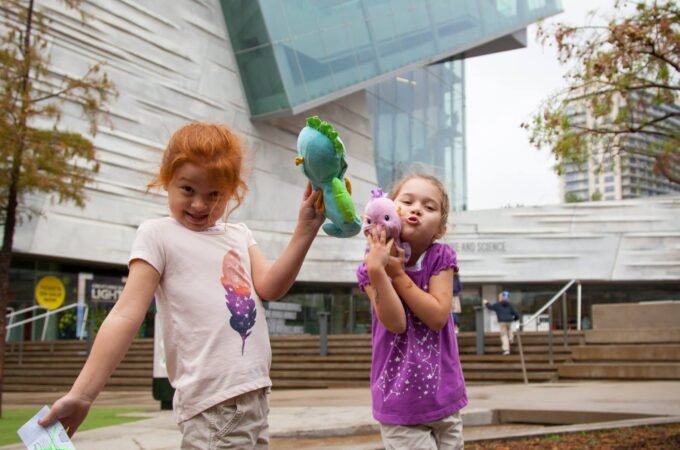 Two children with balloons walking in front of a modern building with a distinctive angular glass structure.