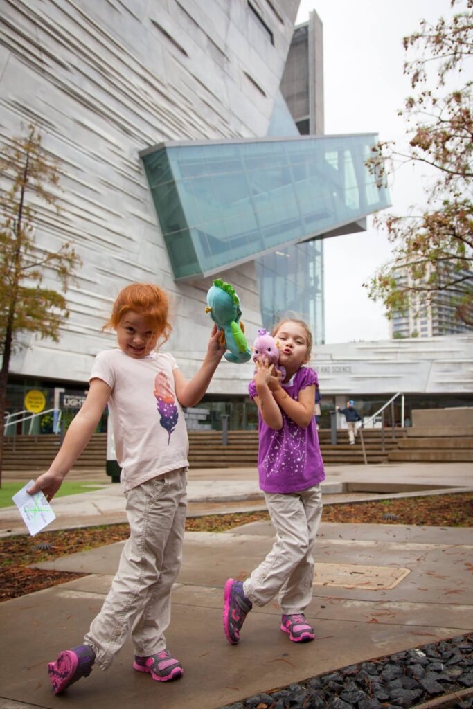 Two children with balloons walking in front of a modern building with a distinctive angular glass structure.