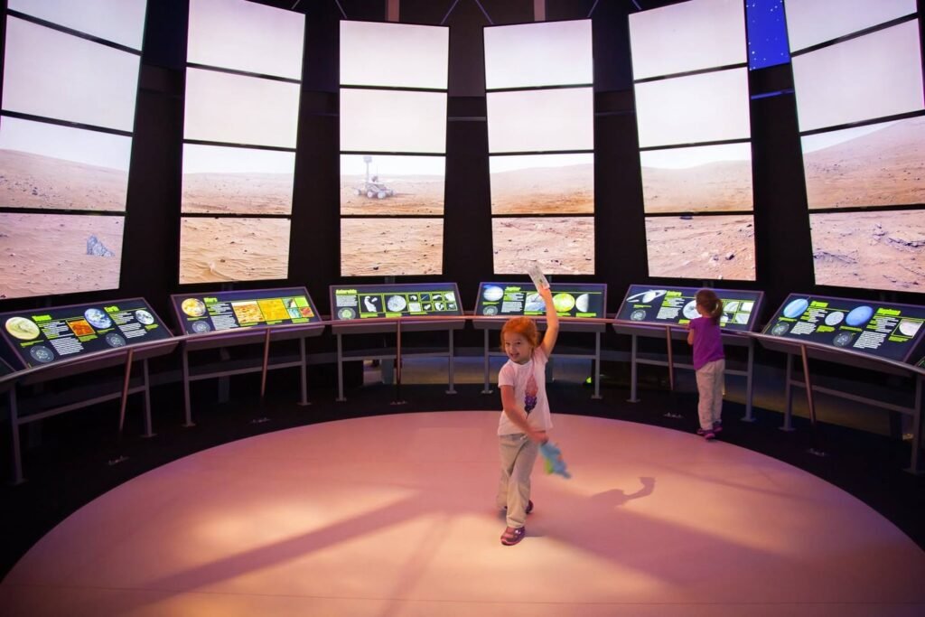 Child in a white tank top standing in a circular interactive planetarium with large screens showing desert landscapes and control panels.