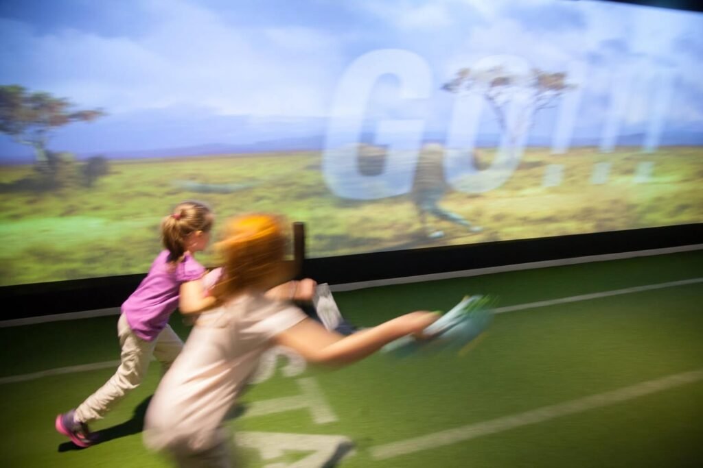 Two children swinging clubs at a drive-thru golf range with a vibrant background wall.