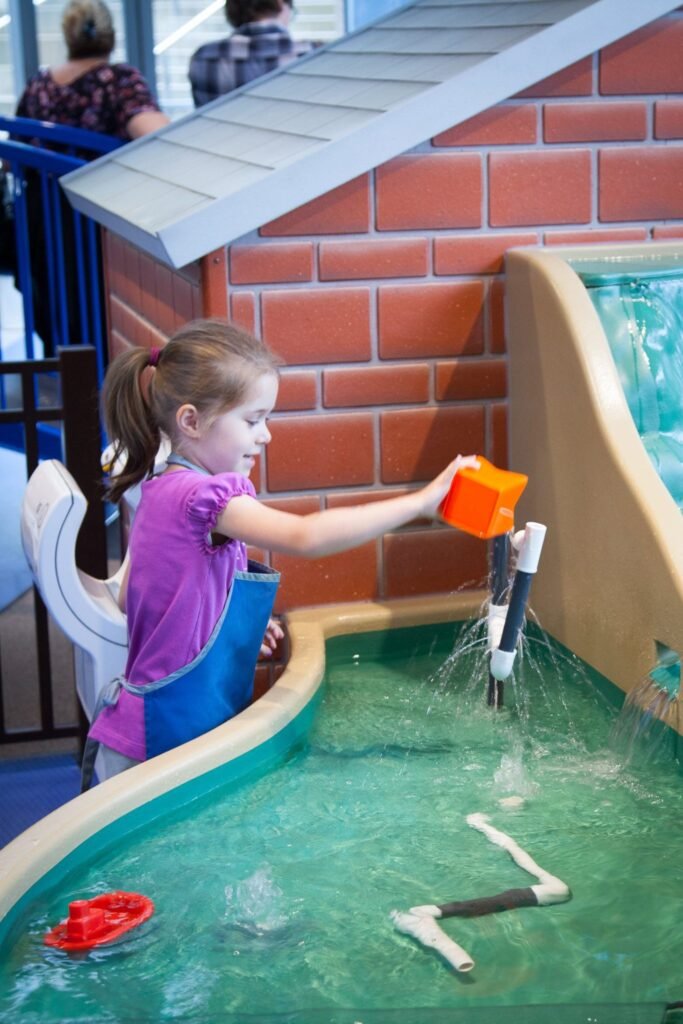 A child in a purple shirt operates an orange lever at a water exhibit, splashing water into a basin.