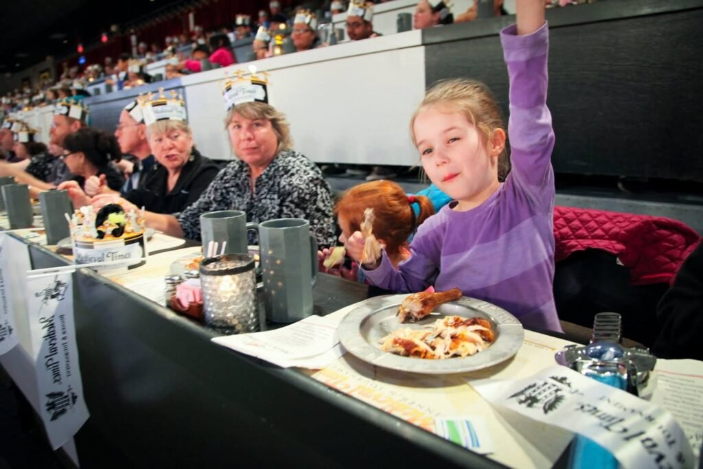 Group seated at a long banquet table with plates and drinks, cheering in stadium-style seating with spectators visible in the background.