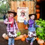 Two children in cowboy outfits at a pumpkin patch with a Wild West wanted poster and a hay bale structure.