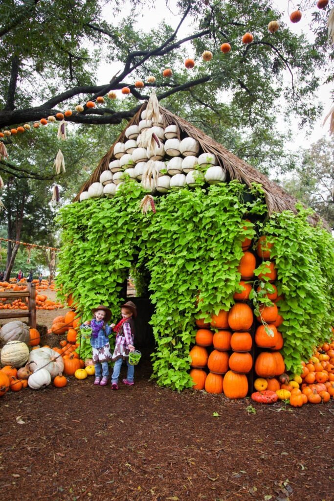 A charming cottage built from stacked pumpkins and gourds covered in green vines, with children in front.