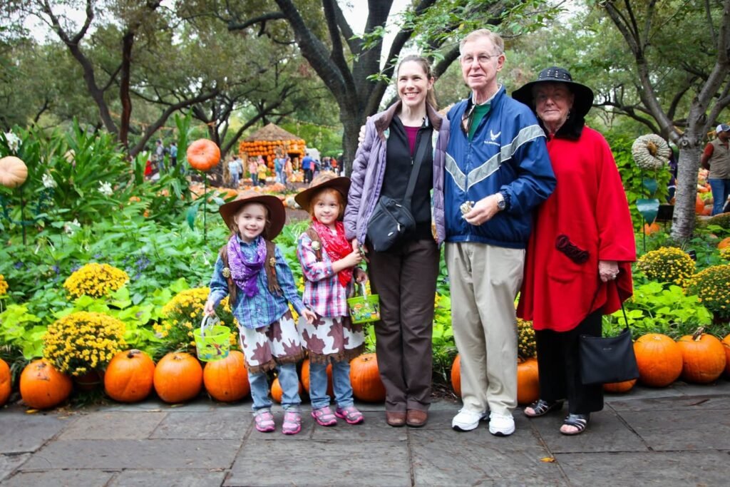 A family enjoys an autumn day at a pumpkin garden, with colorful mums and pumpkins in the background.