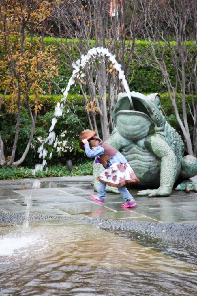 A child holds a sun hat in front of a large green frog statue spouting water in a garden.