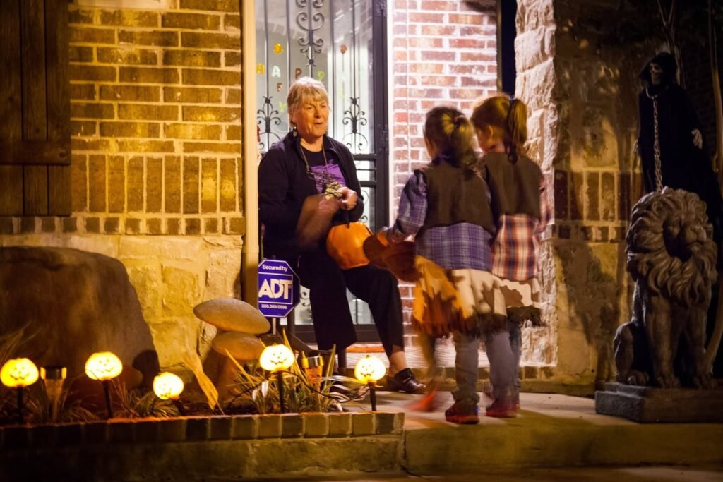 A nighttime scene of a brick building near the water. People are interacting near a statue and illuminated streetlamps along the sidewalk.