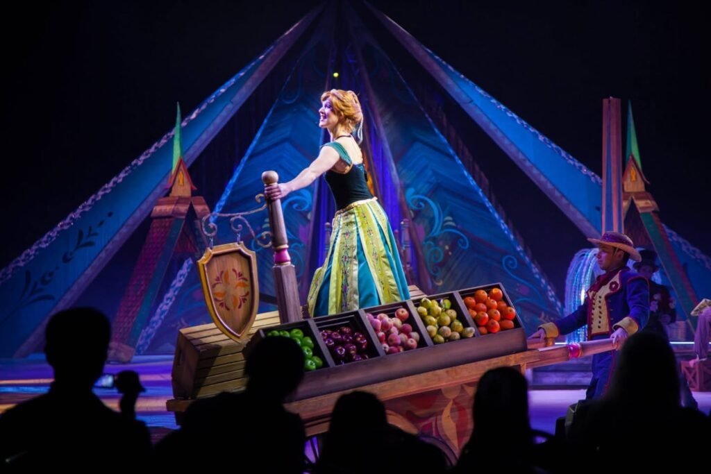 A performer sings at a glowing night boat in Disney World's Enchanted River, surrounded by vibrant stage lights and colorful baskets of fruit.