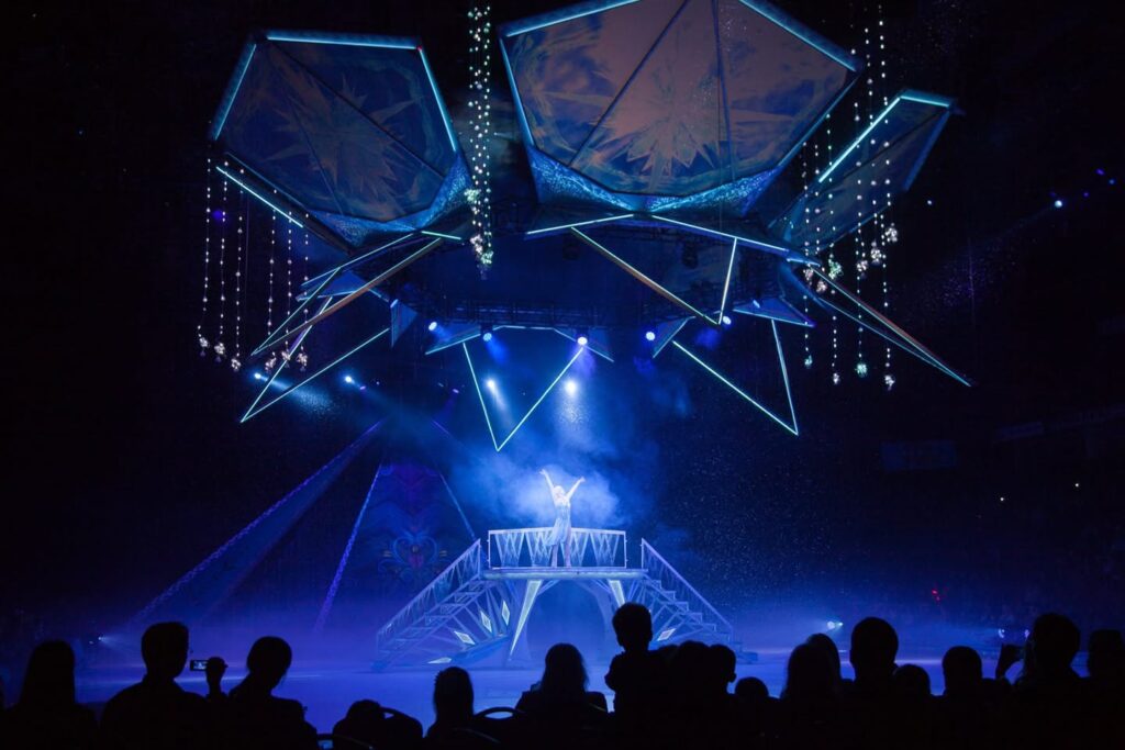 Silhouetted audience viewing a large illuminated stage structure with blue lighting and starburst effects at night.