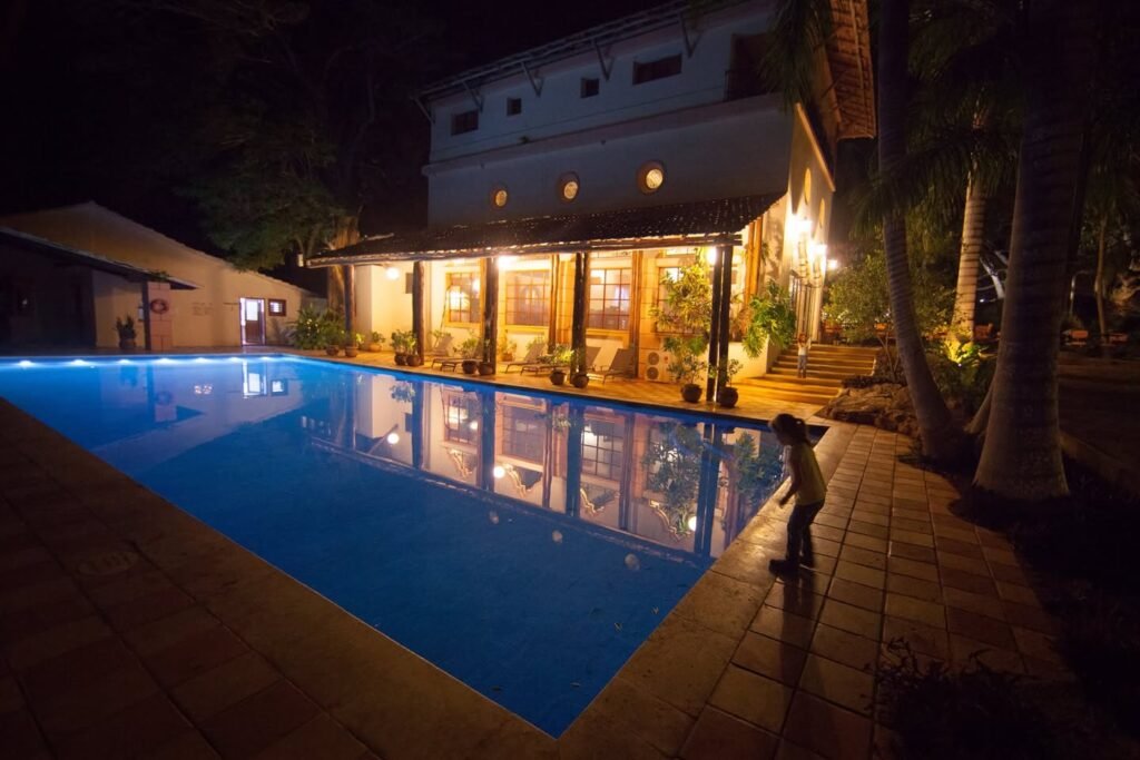 A glowing private swimming pool reflects a warm, lit villa canopy at dusk. Palm trees and tiled decks add to the serene night scene.