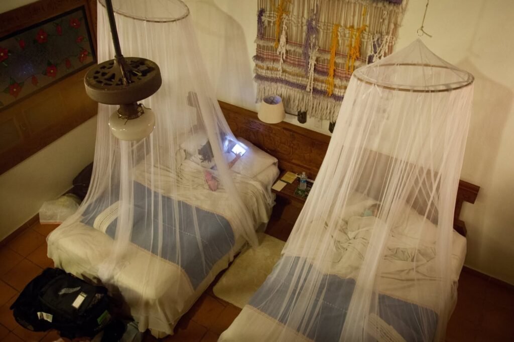 A cozy hotel room featuring two twin beds draped with hanging white mosquito net canopies. The vintage-style setup includes wooden furniture and distinctive decor elements.