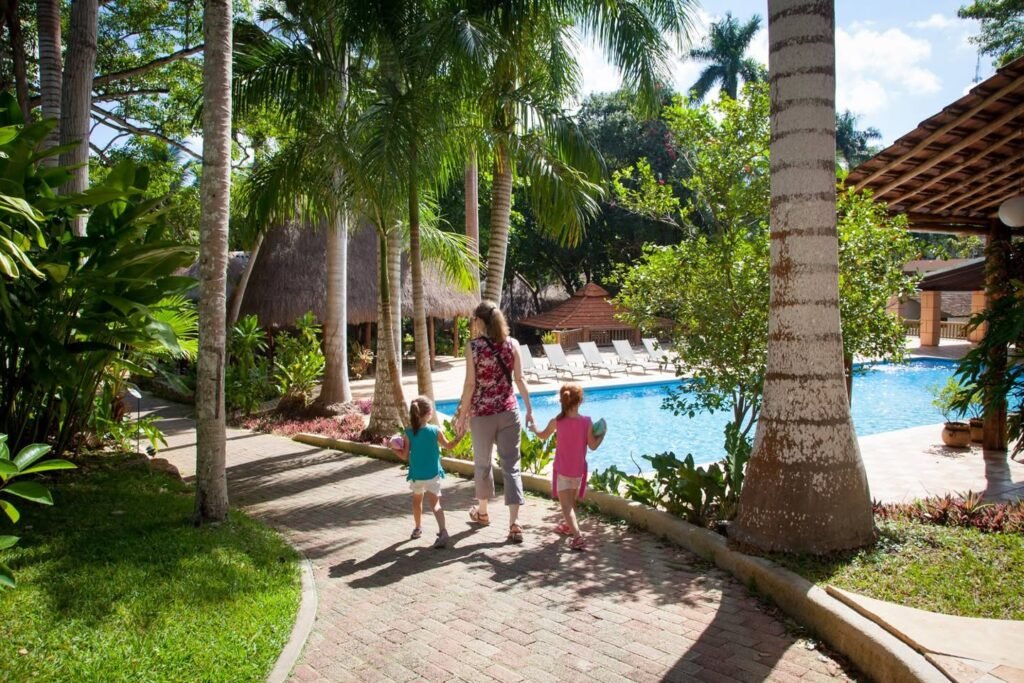 A family strolls along a paved path towards a pool and cabanas, surrounded by tall coconut palms and lush tropical landscaping at a resort villa.
