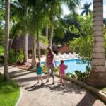 A family strolls along a paved path towards a pool and cabanas, surrounded by tall coconut palms and lush tropical landscaping at a resort villa.