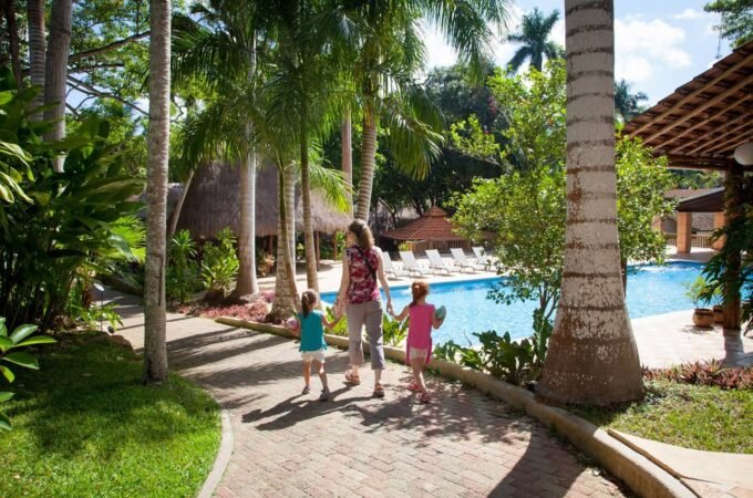 A family strolls along a paved path towards a pool and cabanas, surrounded by tall coconut palms and lush tropical landscaping at a resort villa.
