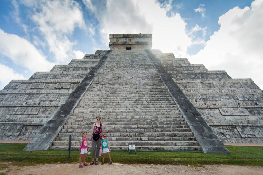 A family stands at the foot of the iconic Chichen Itza pyramid in Mexico. The massive limestone steps rise against a bright, partly cloudy sky.