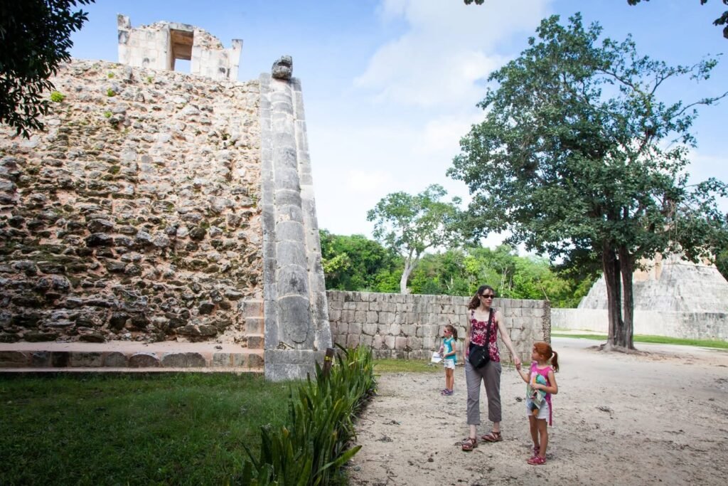 A family explores a Mayan archaeological site with ancient stone structures, a tall balonce top, and surrounding lush green trees under a bright blue sky.