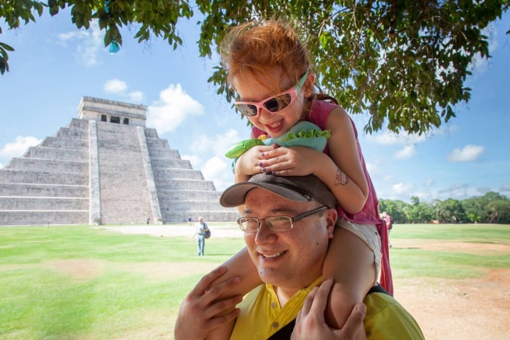 Toddler wearing sunglasses giving piggyback ride with view of El Castillo pyramid at Chichén Itzá on sunny day in Mexico