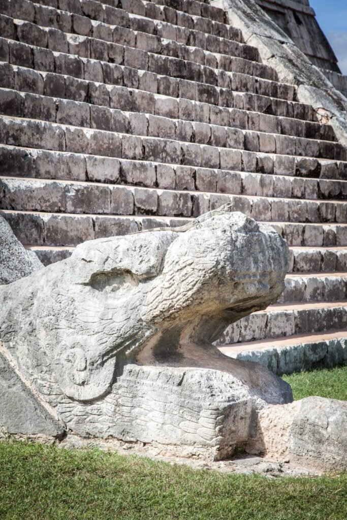 A weathered stone snake head sculpture at the base of the great staircase at Chichen Itza, with close-up details of ancient limestone steps.