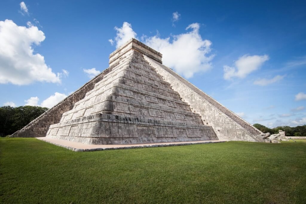 A wide-angle view of the El Castillo pyramid in Chichen Itza, Mexico, showing its stone steps and temple against a bright blue sky with clouds.