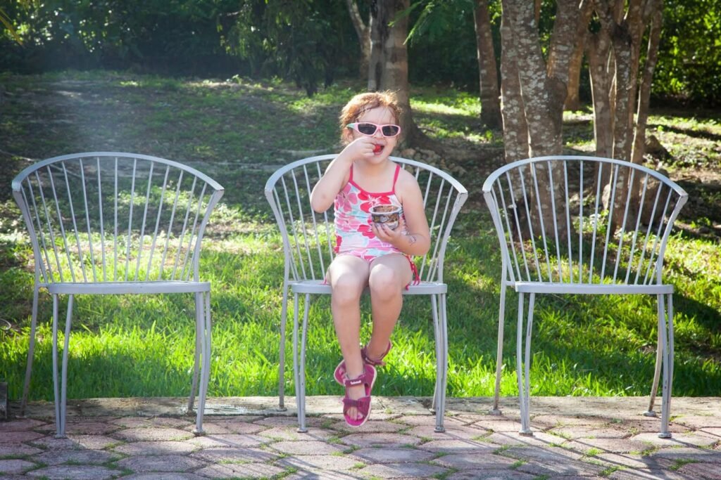 A person wearing sunglasses and a pink outfit sitting on a white metal chair, surrounded by greenery and enjoying a sunny day.